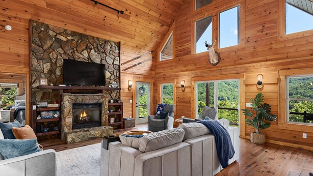 A spacious living room with a stone fireplace, a large TV, and a cozy seating area. - Breathtaking Views! Fast WiFi, Pool Table, Hot Tub, lvl 2 EV Charger, Fire Pit! (Maggie Valley)