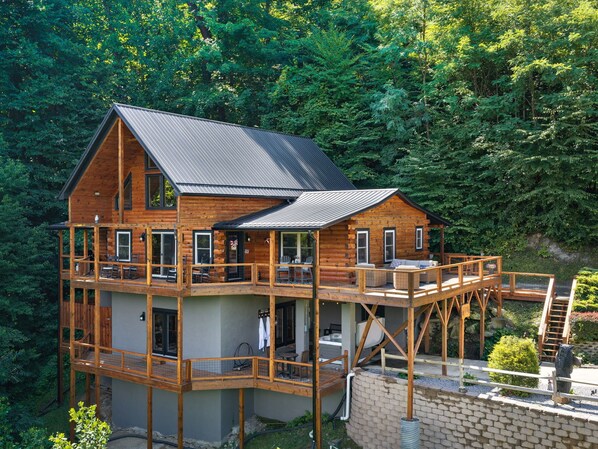 A multi-story log cabin with a balcony, a hot tub, and a view of a forested area. - Breathtaking Views! Fast WiFi, Pool Table, Hot Tub, lvl 2 EV Charger, Fire Pit! (Maggie Valley)