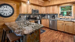 A kitchen with wooden cabinets, a granite countertop, a stainless steel microwave, and a large wall clock. - Breathtaking Views! Fast WiFi, Pool Table, Hot Tub, lvl 2 EV Charger, Fire Pit! (Maggie Valley)