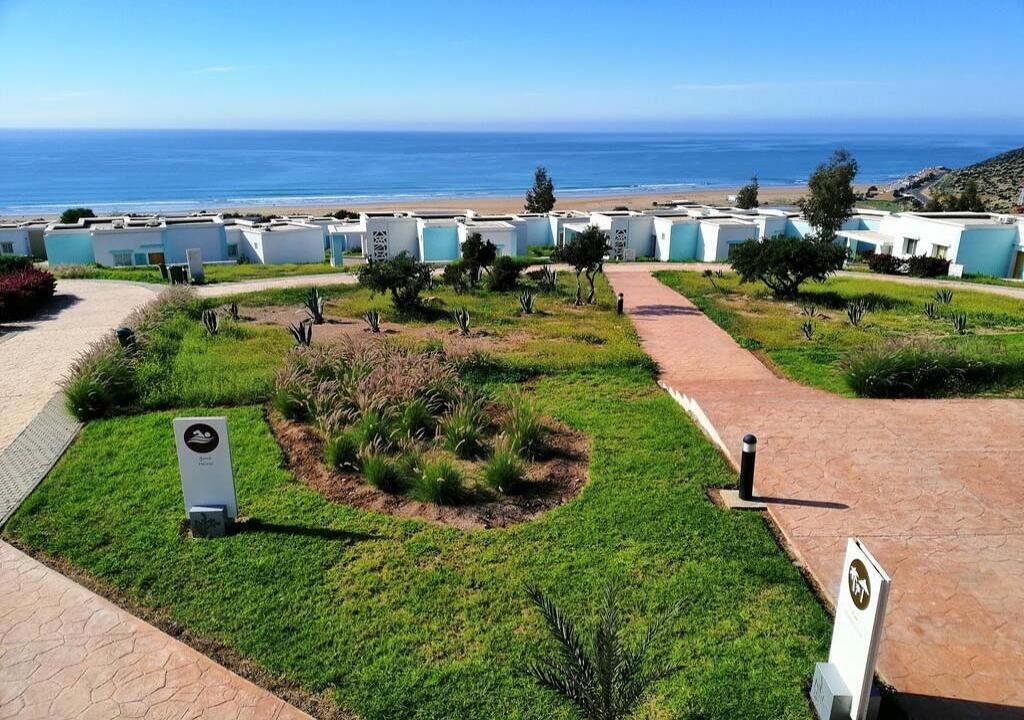 A coastal hotel with multiple white buildings, a paved walkway, and a view of the ocean.