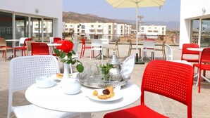 Eine Dachterrasse mit runden Tischen und roten Stühlen, mit einem Tisch gedeckt mit Teekanne, Tassen und einer Auswahl an Gebäck.