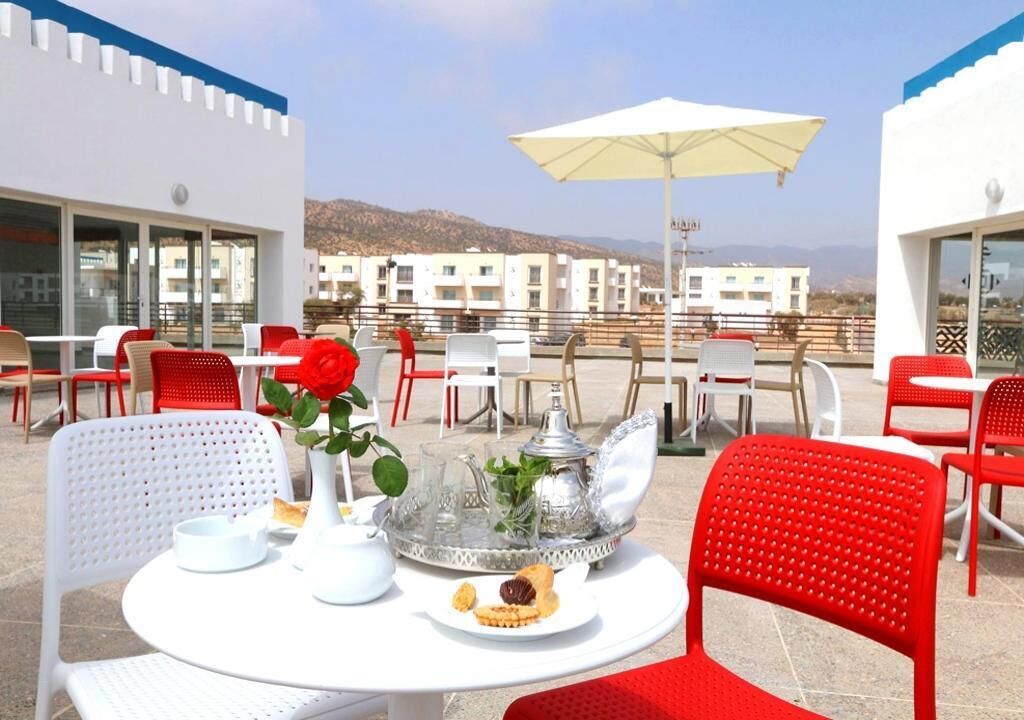 Une terrasse sur le toit, dotée de tables rondes et de chaises rouges, où l’on peut admirer une table dressée avec une théière, des tasses et une assiette de pâtisseries.