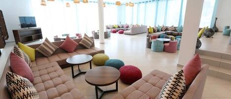 A spacious lounge area with multiple seating options, including sofas and round tables, and a television on the left.