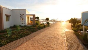A paved pathway leading to a building with a geometric facade and a pool area in the background.