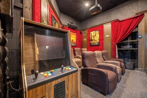 Una habitación con un juego de arcade de madera, sillones reclinables cómodos, una pantalla de proyector y una cortina roja.