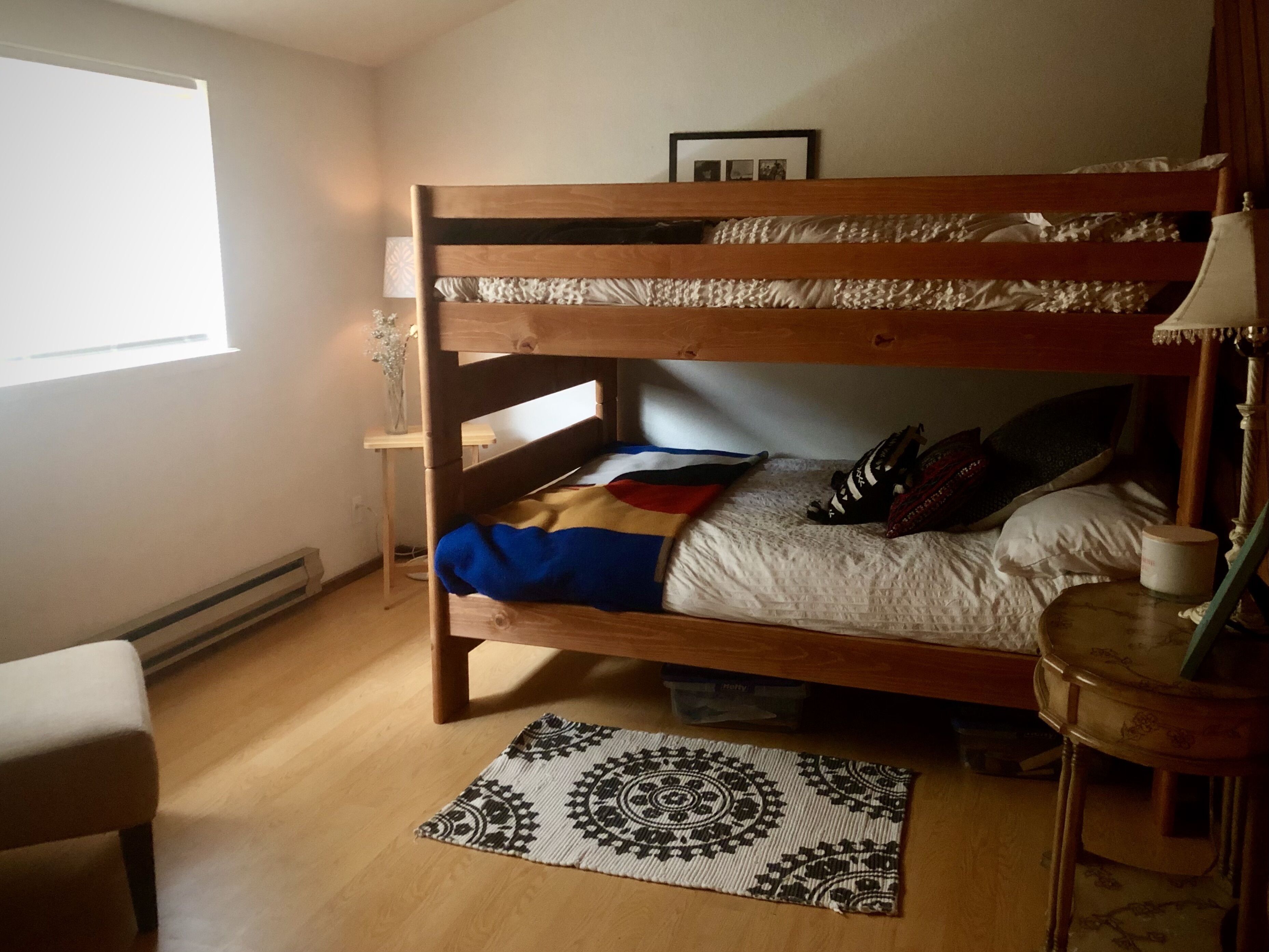 A bunk bed room with a wooden headboard, a bedside table with a lamp, a patterned rug, and a window providing natural light.