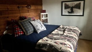 Ein Schlafzimmer mit hölzernem Kopfteil, einem Bett mit gemusterten Betttextilien, einem Nachttisch mit Lampe und einem gerahmten Bild an der Wand.