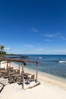 Una playa con mesas y sillas de madera bajo una estructura de madera, con vista al océano.