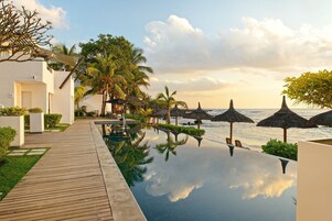 Un área de piscina tranquila con terraza de madera, palmeras y sombrillas de paja con vistas al océano.