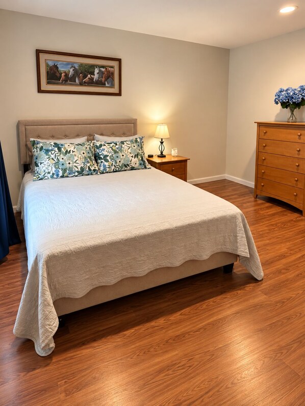 Un dormitorio con cama, mesita de noche, cómoda, lámpara y un cuadro en la pared.