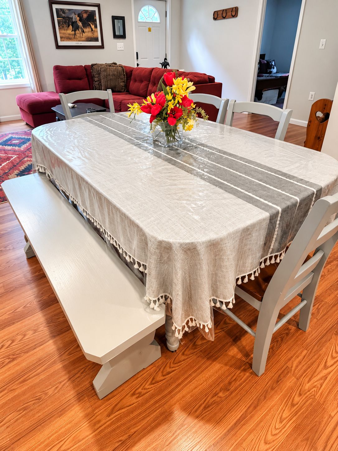 Une table à manger recouverte d’une nappe, des bancs et des chaises, dans une pièce au plancher en bois et avec un canapé rouge en arrière-plan.