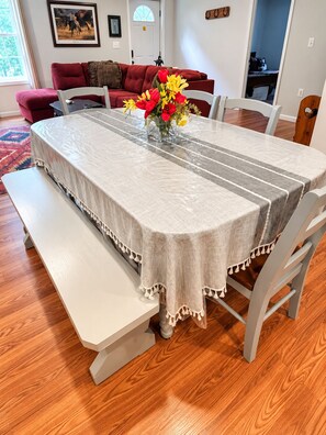 A dining table with a tablecloth, benches, and chairs in a room with wooden flooring and a red sofa in the background. - The Meadow Lodge- Cozy 3BR Farm Barndo with Fire Pit and nearby Wedding Venues  (White Hall)