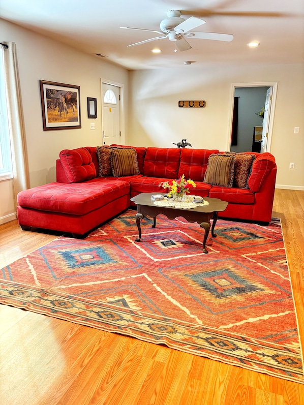 A living room with a red sectional sofa, a coffee table with a vase of flowers, a patterned rug, and a ceiling fan.