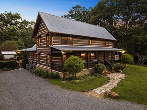A two-story log cabin with a porch, stone foundation, and a gravel driveway. - Waterfall Pool · Barrel Sauna + Hot Tub · Cabin · 15 Private Acres (Mineral Bluff)