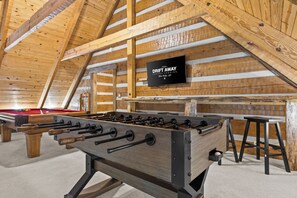 An indoor recreational area with a foosball table, a pool table, and a television screen displaying 'DRIFT AWAY LUXURY LODGING Blue Ridge, Gold'. - Waterfall Pool · Barrel Sauna + Hot Tub · Cabin · 15 Private Acres (Mineral Bluff)