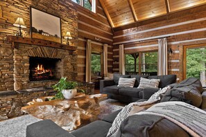 L’intérieur chaleureux d’un chalet avec une cheminée en pierre, un ensemble de canapés et une grande fenêtre donnant sur le paysage extérieur.