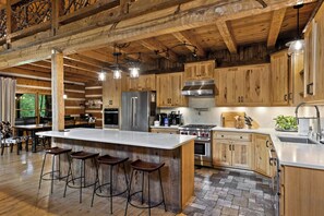 A spacious kitchen with wooden cabinets, a large island with stools, and modern appliances. - Waterfall Pool · Barrel Sauna + Hot Tub · Cabin · 15 Private Acres (Mineral Bluff)