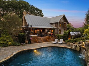 A log cabin with a swimming pool, outdoor seating, and a stone fireplace.