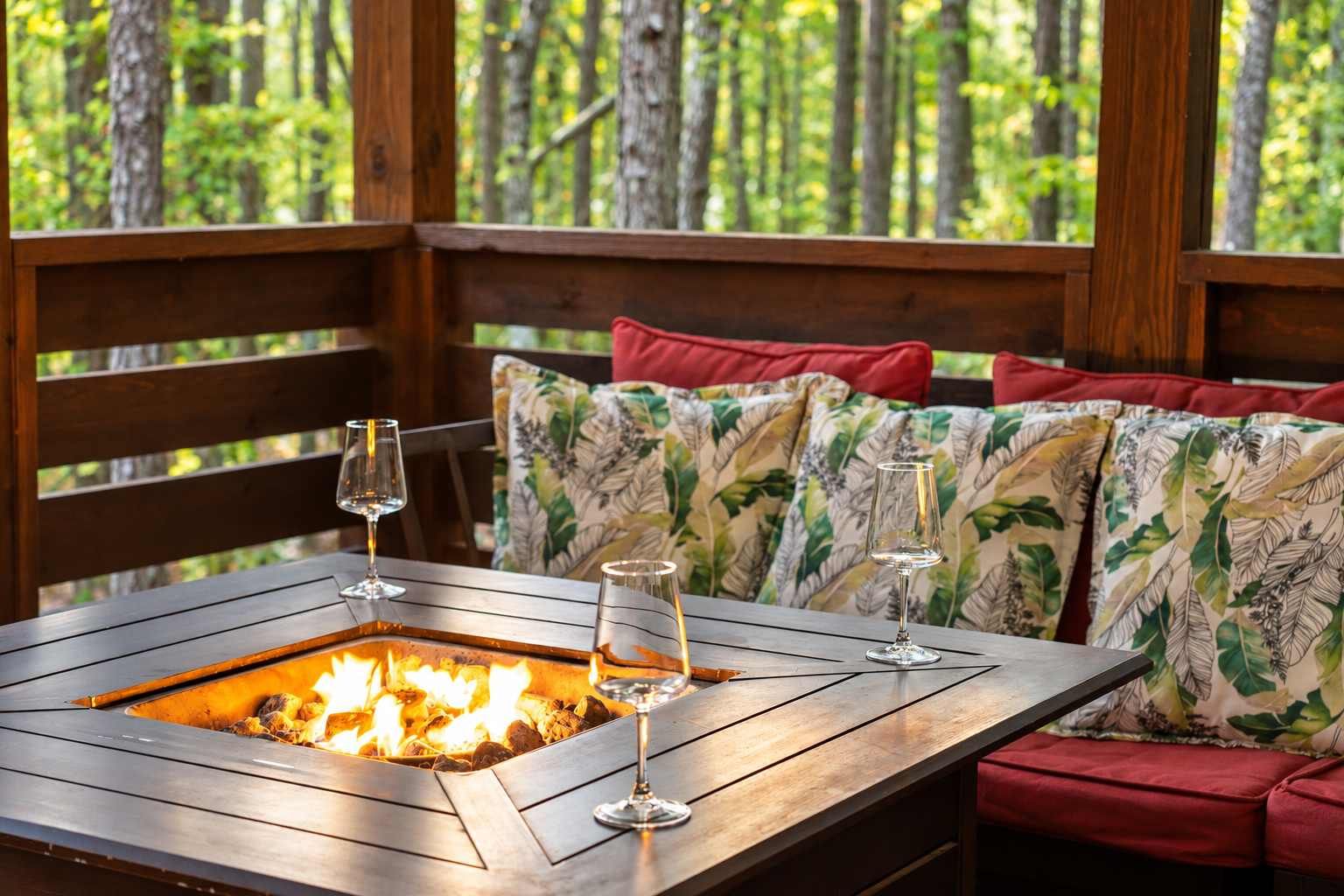 A wooden deck with a built-in fire feature, cushioned seating with patterned cushions, and a glass table with wine glasses.