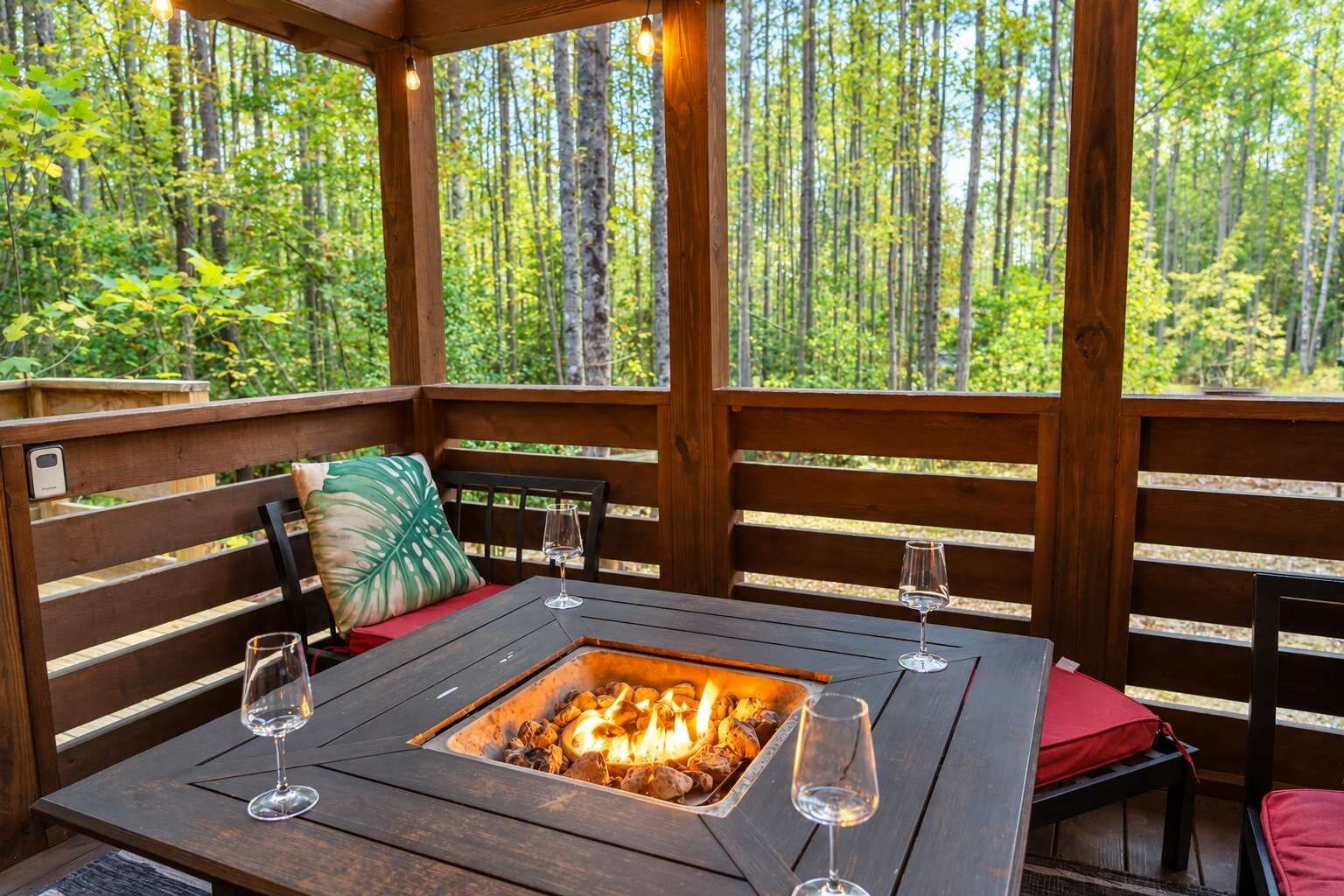 Eine Terrasse aus Holz mit Feuerkorb-Tisch, zwei Stühlen und einer Bank mit Kissen.