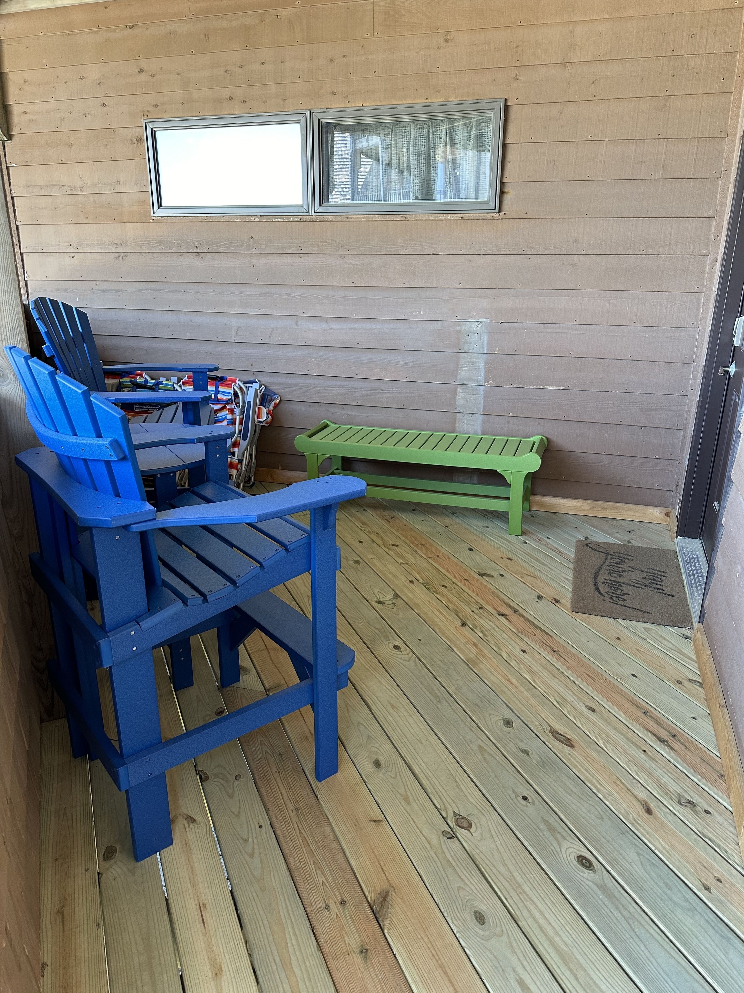 A wooden deck with blue and green Adirondack chairs, a small table, and a window with blinds.