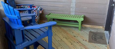 A wooden deck with blue and green Adirondack chairs, a small table, and a window with blinds.
