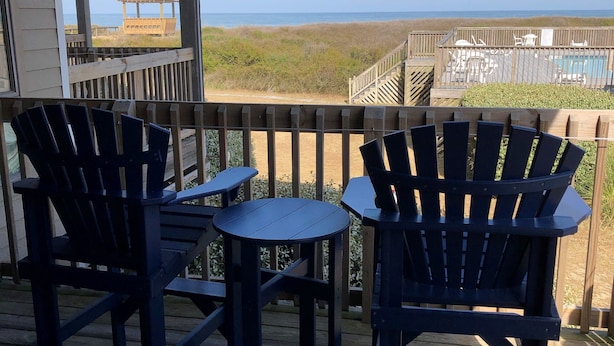 Eine Holzterrasse mit zwei Adirondack-Stühlen und einem kleinen Tisch, mit Blick auf einen Strand und einen Poolbereich.