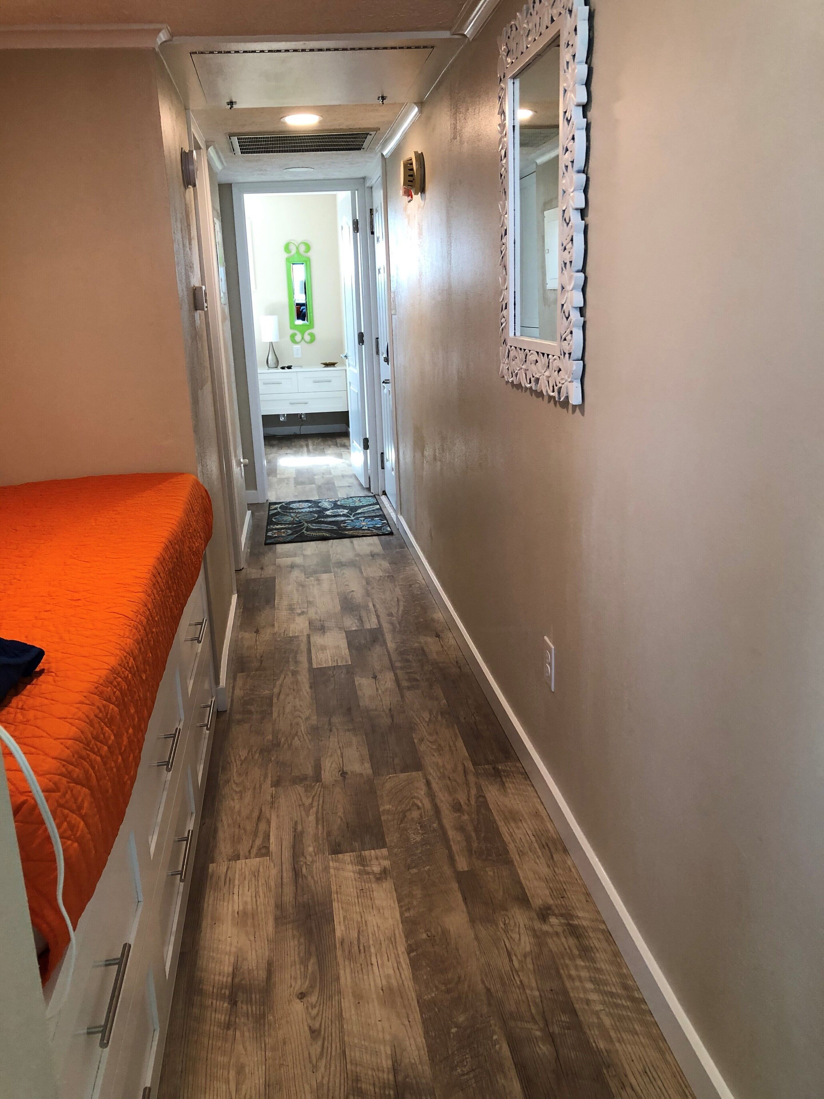 A narrow hallway with a wooden floor, a bed with an orange cover, and a mirror on the wall.