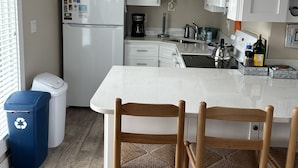 Eine Küche mit weißen Schränken, einem Kühlschrank, einer Kaffeemaschine und einem Spülbecken. Zwei Korbstühle stehen an der Kücheninsel.