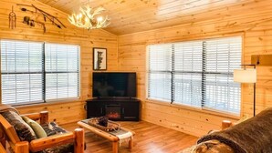 A log cabin interior with a fireplace, wooden furniture, and large windows.