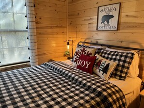 A cozy bedroom with a checkered bedspread, a pillow with 'JOY', a wall-mounted light, and a bear-themed poster. - Osito Cabin, Cozy Escape w/ Hot Tub and Firepit in the Heart of Broken Bow (Broken Bow)
