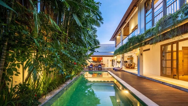 A pool with a wooden deck, surrounded by lush greenery and a building with large windows and a balcony. - Fabulous Private Villa, 3 BR, Canggu With Staff (Tibubeneng)