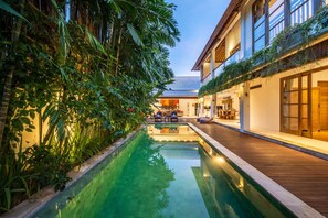A pool with a wooden deck, surrounded by lush greenery and a building with large windows and a balcony. - Fabulous Private Villa, 3 BR, Canggu With Staff (Tibubeneng)