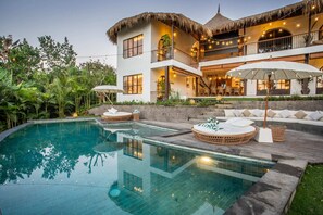 A modern house with a large swimming pool, outdoor seating, and a thatched-roof balcony.