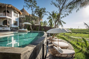 A poolside area with lounge chairs, a thatched-roof building, and a view of a green landscape.