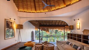 A spacious room with a large bed, a sofa, and a dining table. There is a view of a lush garden through an arched window.