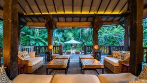 A covered outdoor seating area with wicker furniture and a central umbrella, surrounded by lush greenery.