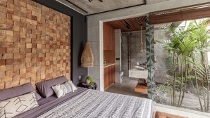 A modern bedroom with a wooden wall, a bed with patterned bedding, a small bedside table, and a view of a garden through a glass door. - Batukaru Private Pool Suite 1 (Canggu)