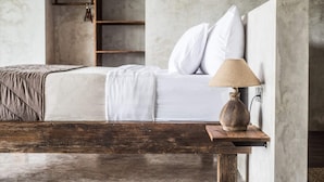 A bedroom with a bed, white bedding, a wooden nightstand with a lamp, and a concrete wall. - Exceptional 2 BR Suites in Ubud Hidden Gem (Ubud)