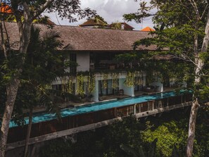A poolside view of a building with a long swimming pool, surrounded by lush greenery and trees.