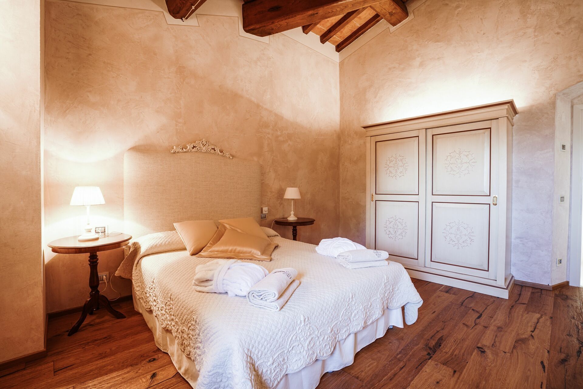 A bedroom with a wooden floor, a bed with white linens, two bedside tables with lamps, and a wardrobe with decorative panels.