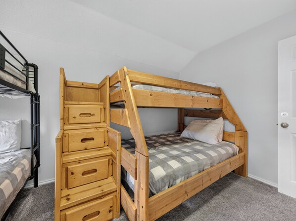 Un lit superposé avec une échelle en bois et des tiroirs, un lit inférieur recouvert d’une couverture à carreaux et une porte blanche.