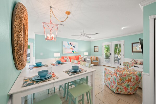 A dining area with a white table, blue plates, and green chairs. There is a ceiling fan, a woven wall hanging, and a colorful painting on the wall.