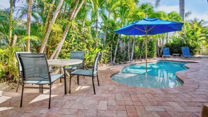 A poolside area with a table and chairs, surrounded by lush greenery and palm trees.