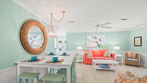 A dining area with a white table, teal chairs, and a woven mirror. There is a ceiling fan and a painting on the wall.