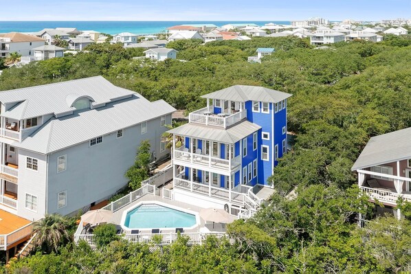 A large house with a swimming pool, surrounded by dense greenery and a view of the ocean.