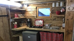 Ett kompakt kök med en spis, diskho och ett kylskåp. Det finns hyllor i trä där köksredskap förvaras, och ett fönster ovanför diskhon.