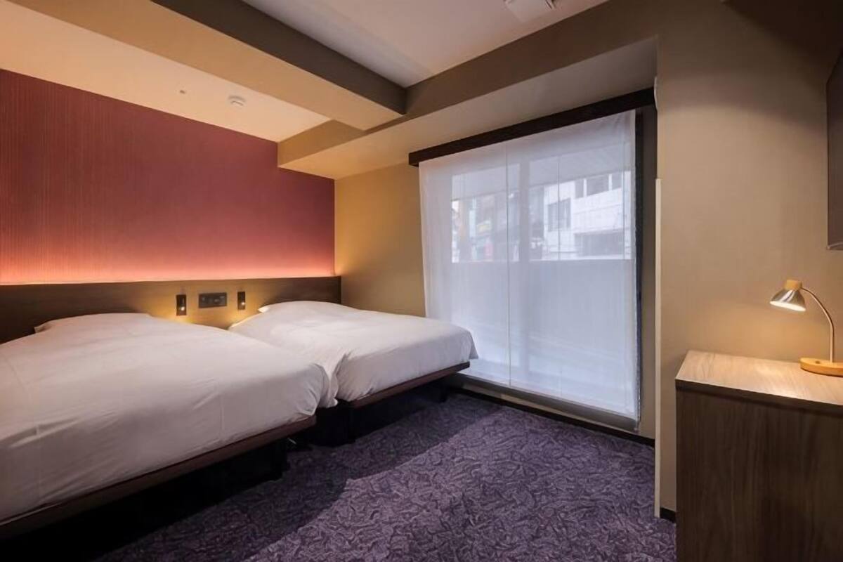 A hotel room with two beds, a bedside table, a window with blinds, and a wall-mounted light fixture.