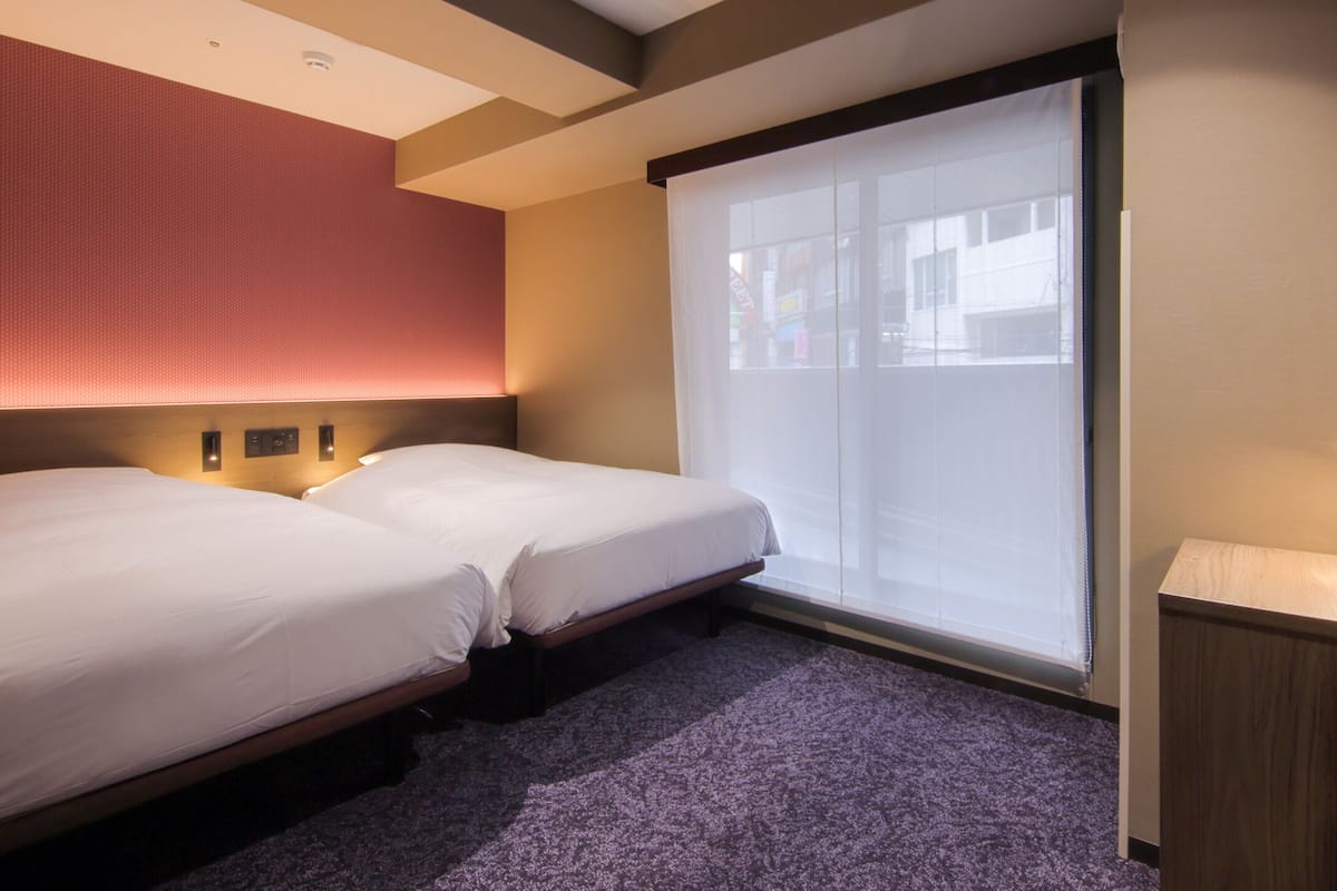 A hotel room with two single beds, a large window with blinds, a small desk with a lamp, and a wall-mounted TV.