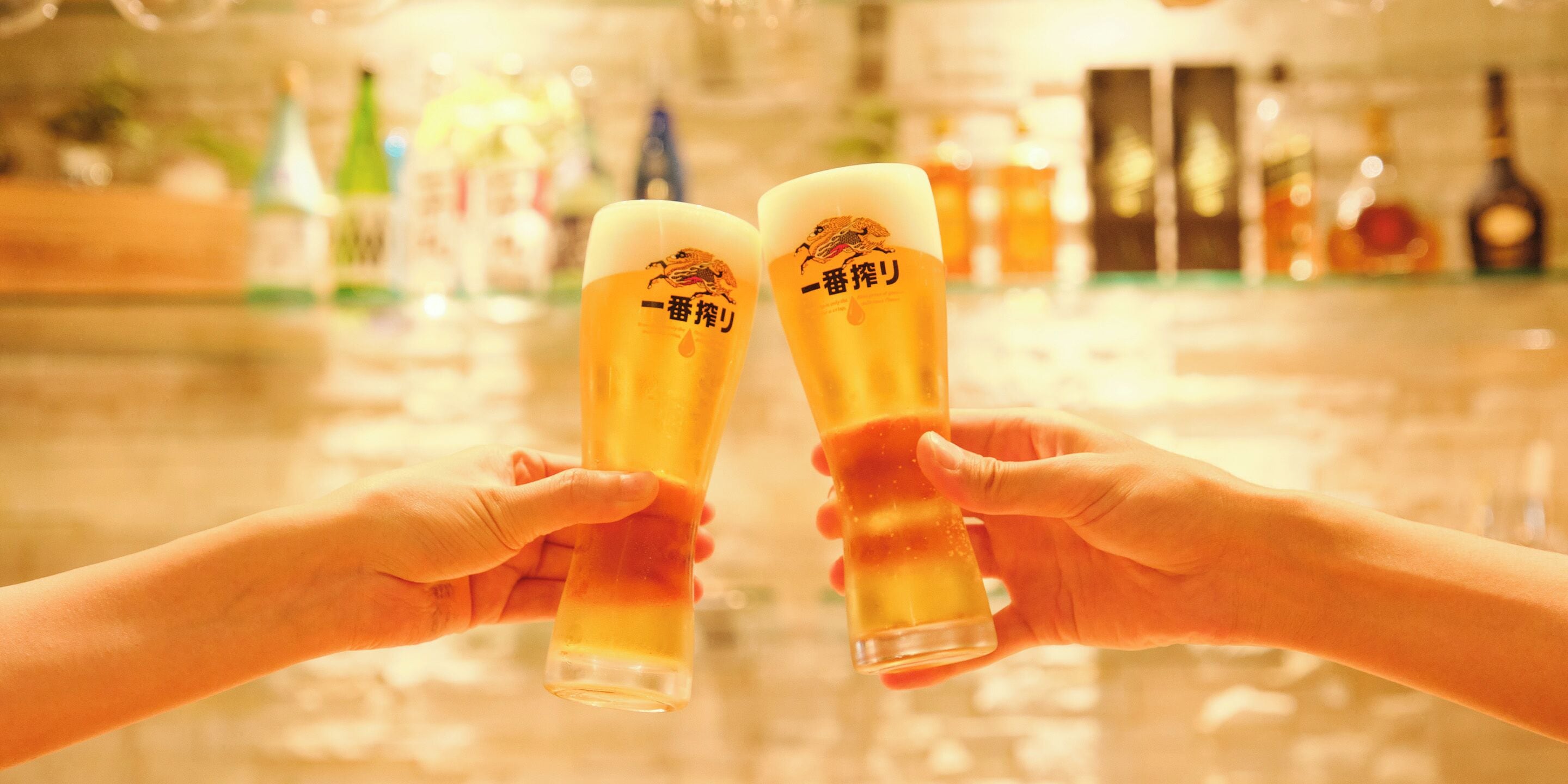 Two hands holding a tall glass with a logo, in a bar setting with bottles in the background.